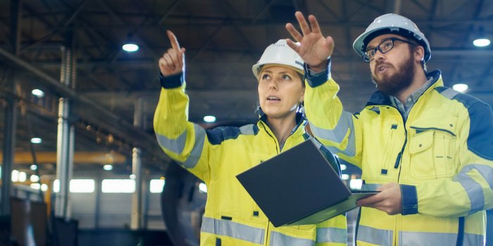 Eine Frau und ein Mann in einer Warnschutzjacke und einem Bauhelm in einer Halle