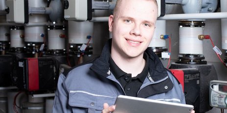 Ein junger Anlagenmechaniker vor einer Heizungsanlage, der ein Tablet hält 